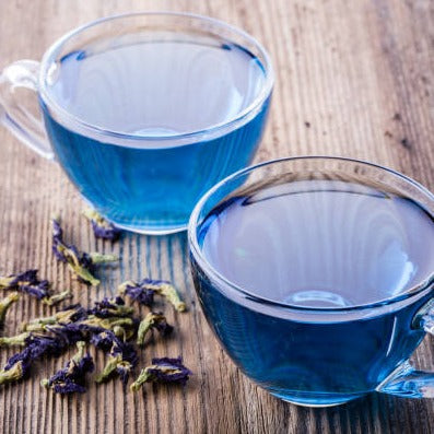 Two glass cups of vibrant blue butterfly pea flower tea with dried petals on wooden table.